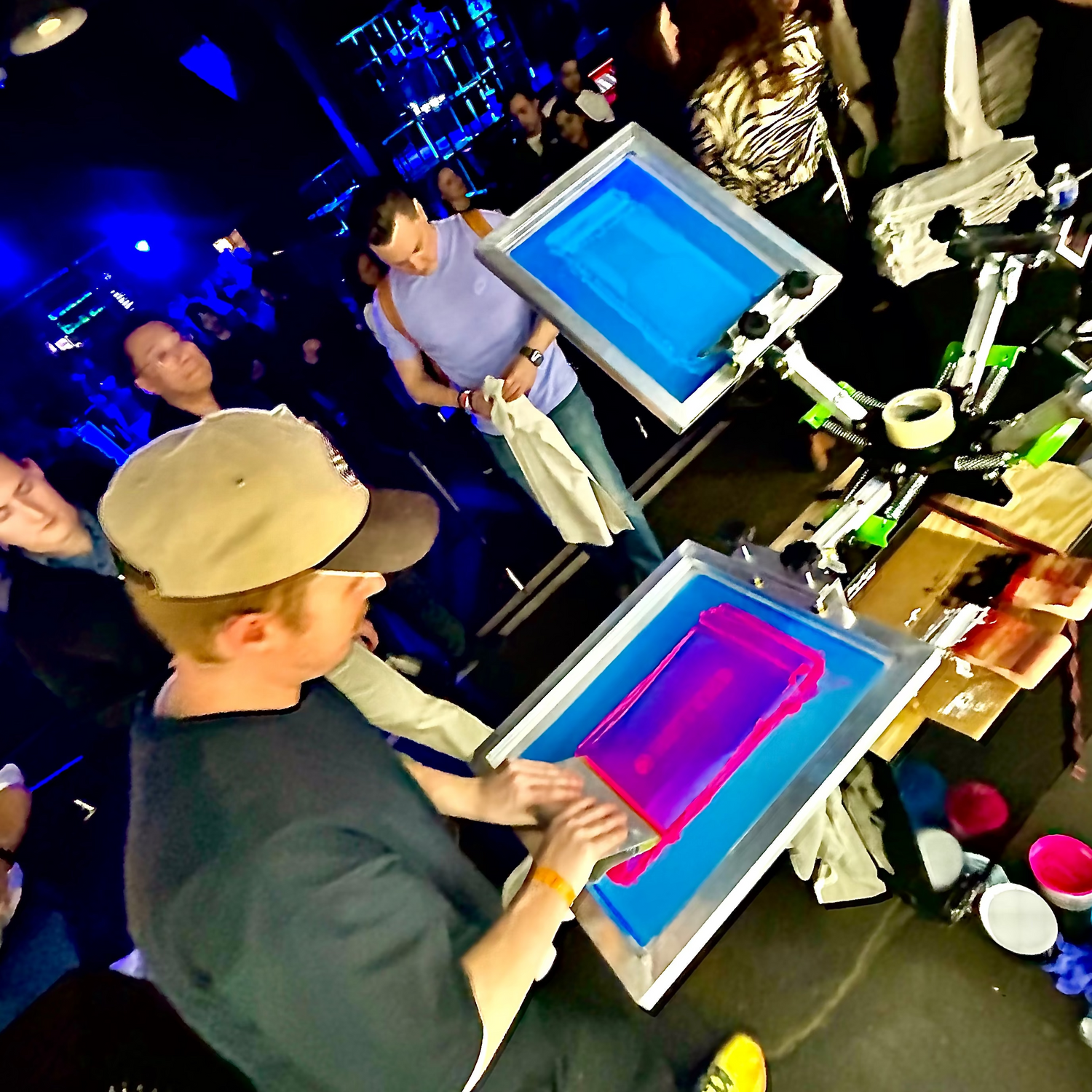 A group of happy event goers are gathered around a onsite printing technician at a Live Screen Printing, Neon Print Party where the color of neon inks a glowing from black lights illuminating the room.