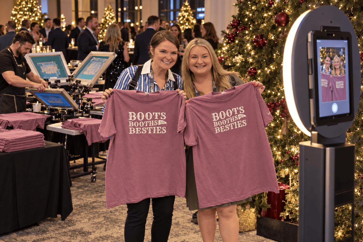Two women at a corporate holiday Christmas event holding up t-shirts that were live screen printed and posing for a photo booth. The background features a decorated Christmas tree, a live screen printing kiosk, and a bar with people enjoying the holiday party.