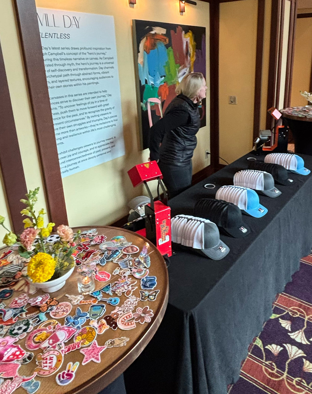 A live hat bar event at a convention in Mandalay Bay Conference Center in Las Vegas with baseball caps and trucker hats, A hat technician stand in the back ground and a round wooden table in the foreground is covered in colorful custom patches.