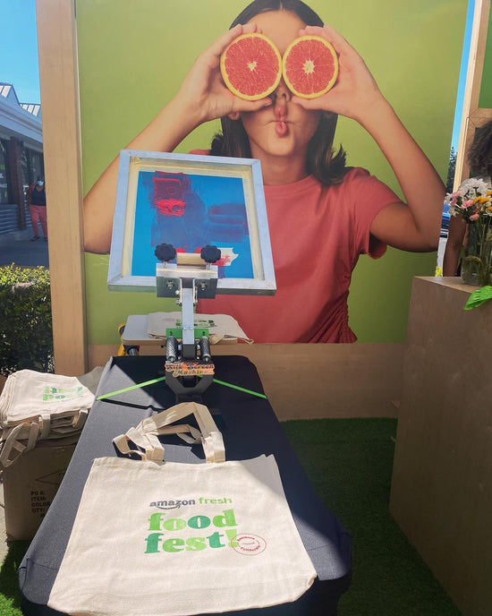 Experitintial Marketing booth for Amazon company with a person holding sliced grapefruits over eyes with a display of a live screen printing press and 'Amazon Fresh Food Fest' tote bag.