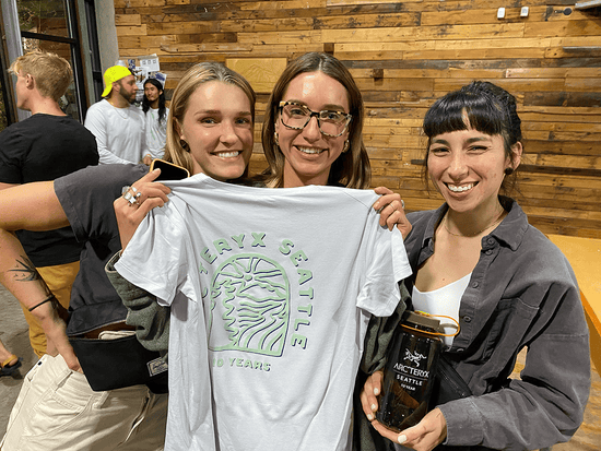 Three women at a live screen printing event holding a t-shirt with the Arc'teryx logo freshly printed on it in Seattle, WA.