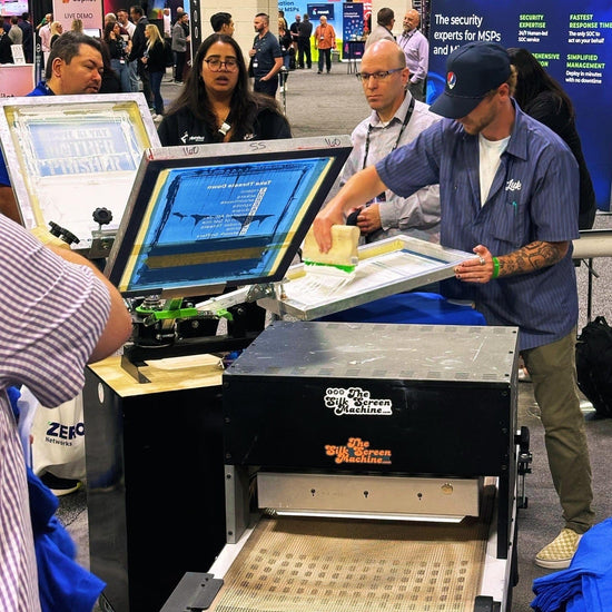 A crowd at a corporate conference gathered around a professional screen printer who is live screen printing, onsite, at an event in a trade show booth.