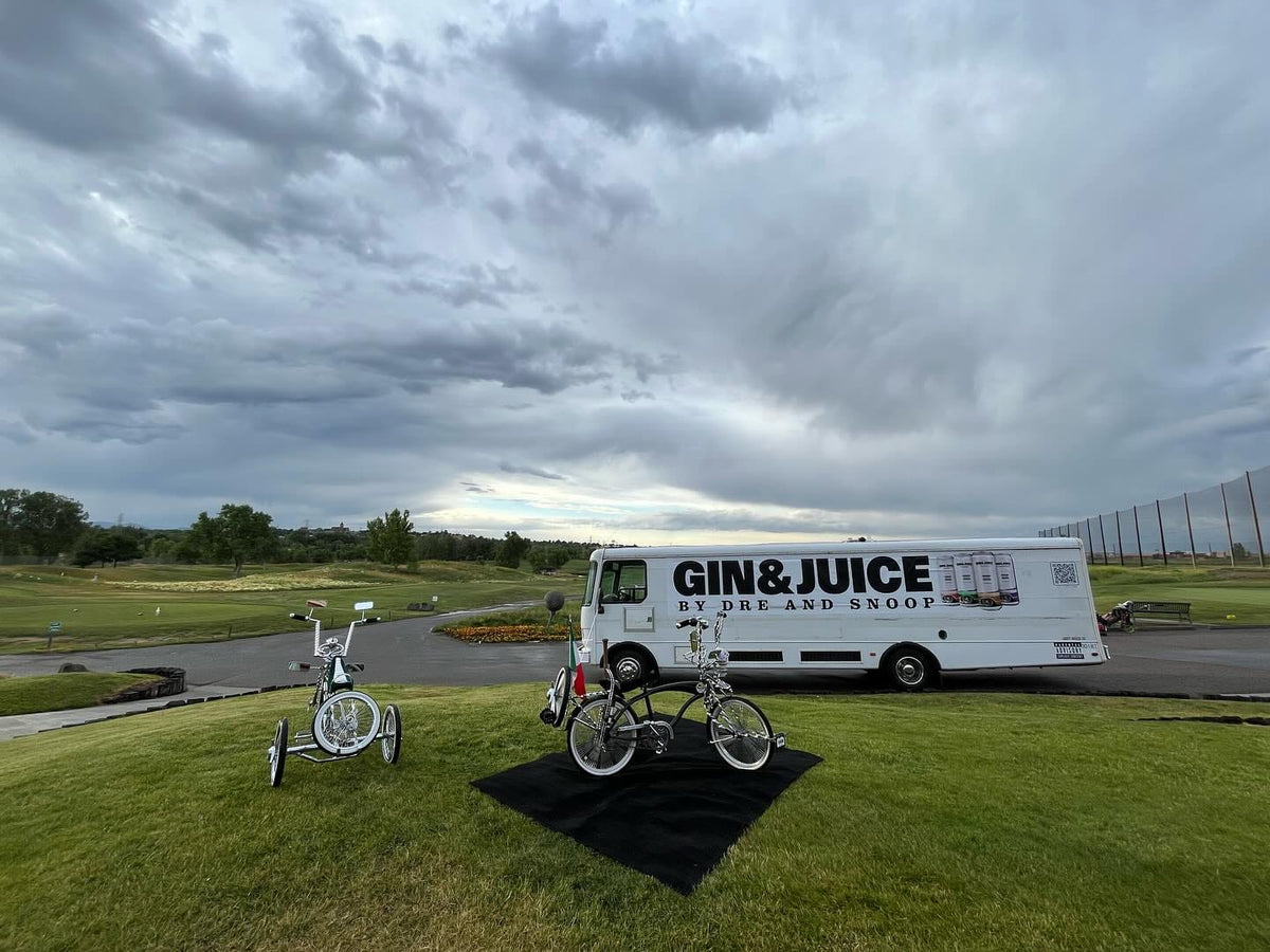 Van labeled 'GIN&JuICE' with two low rider bicycles on a grassy area under a cloudy sky for a mobile billboard campaign in Denver, Colorado.