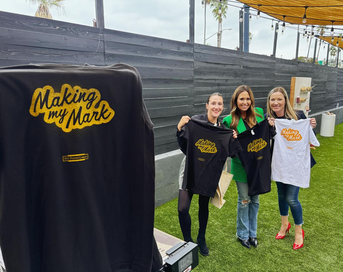 Three women at a Fortune 500 company live onsite screen printing event holding up t-shirts with a yellow 'Making my Mark' logo, next screen printing press. Live Customizing Promotional Products in Los Angeles, California. 