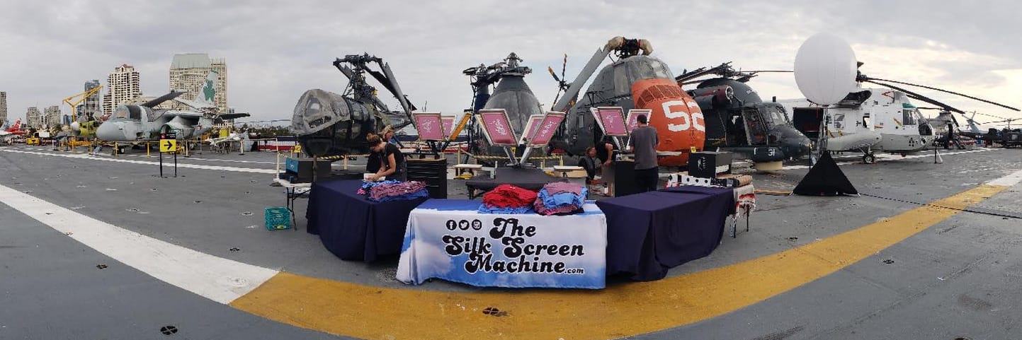 A custom blue and white banner reading 'The Silk Screen Machine' displayed on a table for a VIP live screen printing event on the deck of a military aircraft carrier, the USS Midway, with helicopters and aircraft in the background.